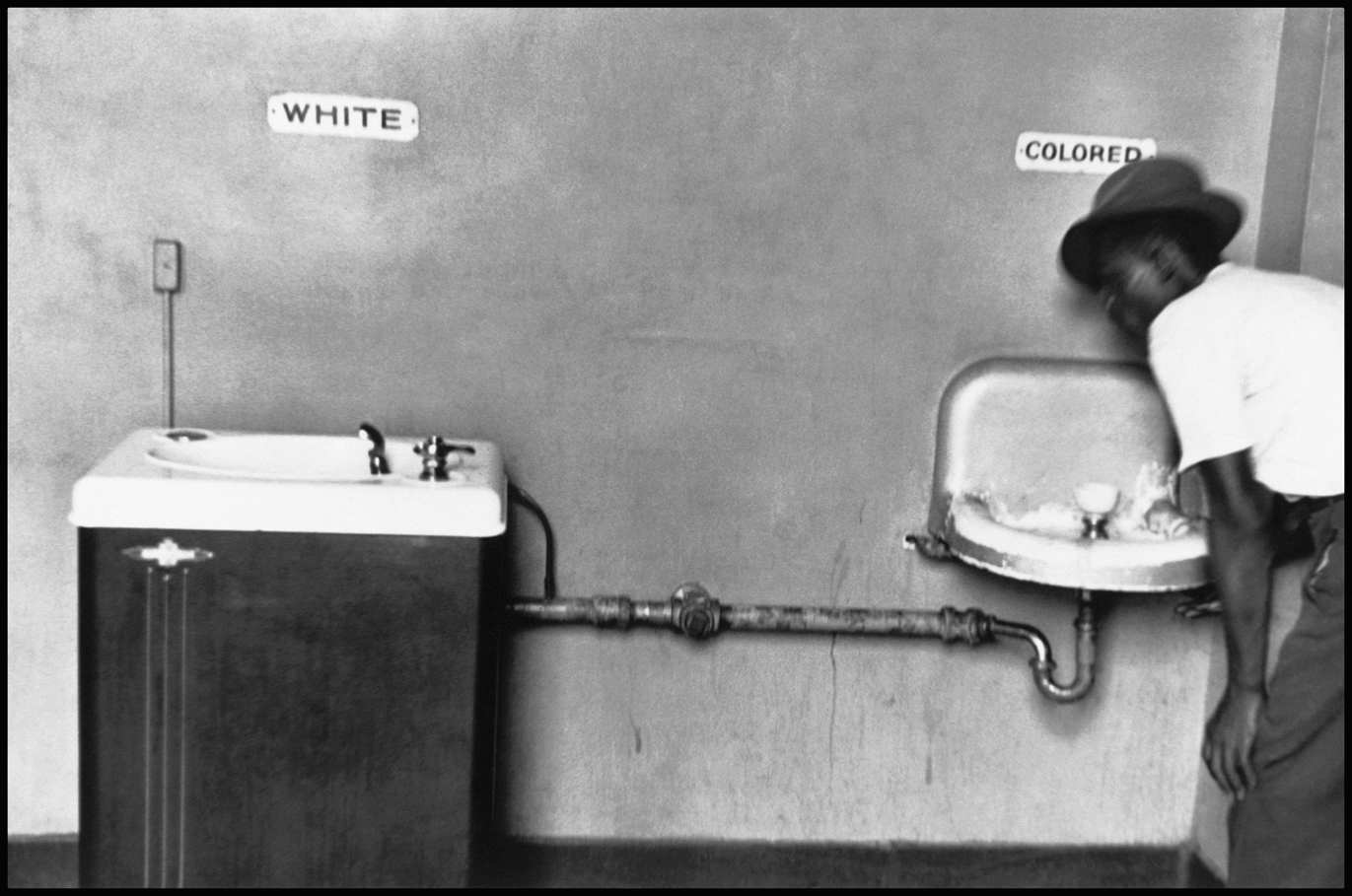 The black and white photo shows two types of water fountains in North Carolina in 1950. On the left is a well-built water fountain which is labelled as “WHITE,” whereas on the right there is another one which is not properly built and looks like a primitive wash basin. The latter is labelled as “COLORED.” A black man in pant-shirt, wearing a hat, is bent forward to drink water from the water fountain.