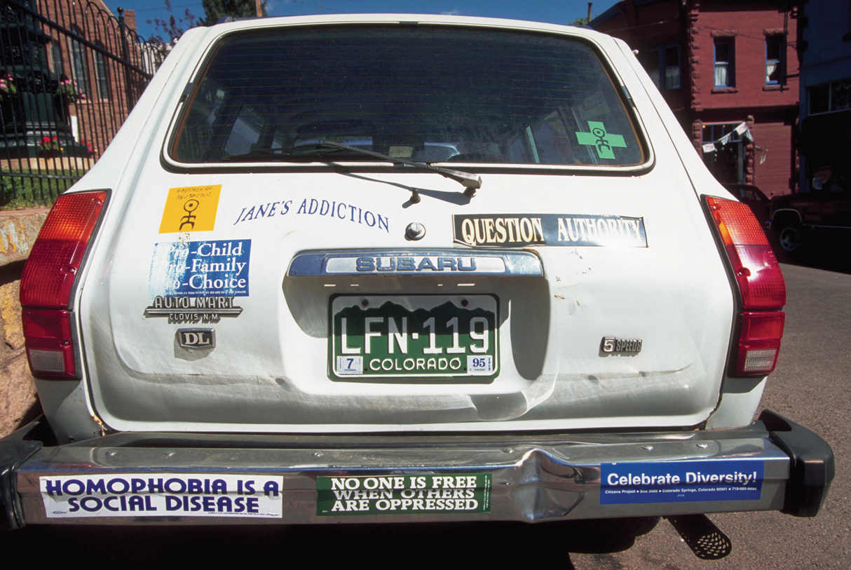 In the photograph, the back side of a hatchback car is shown with bumper stickers on it, one sticker on the bumper reads, ‘Homophobia is a social disease’. Another sticker reads, ‘No one is free when others are oppressed’. Yet another sticker reads, ‘Celebrate Diversity’. Other stickers read, ‘Jane’s Addiction’, and ‘Question Authority’. There is a plus symbol also, on which a figure is drawn. The license plate of the car reads, ‘LFN-119’, and below the number, it reads, ‘Colorado’.