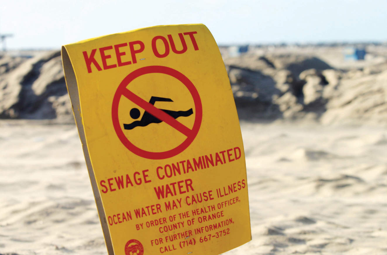 A yellow sign board warning says ‘KEEP OUT, SEWAGE CONTAMINATED WATER, OCEAN WATER MAY CAUSE ILLNESS, By order of the Health Officer, County of Orange, For further information, Call (714)667-3752’. The warning also shows a symbol of No Swimming.