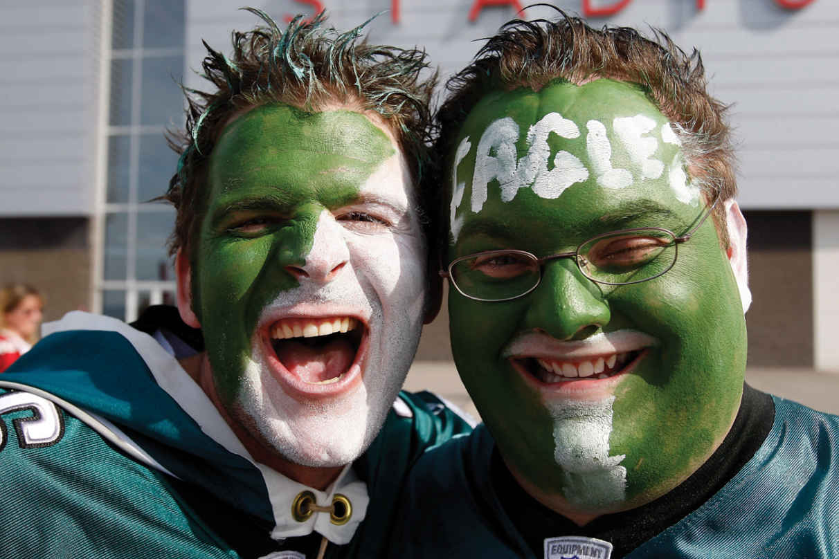 A photograph of two men who have painted their faces in green and white, laughing and looking cheerful marked as ‘Philadelphia Eagles’ fans by an arrow. The right-hand side man also has ‘EAGLES’ written in white color on his forehead.