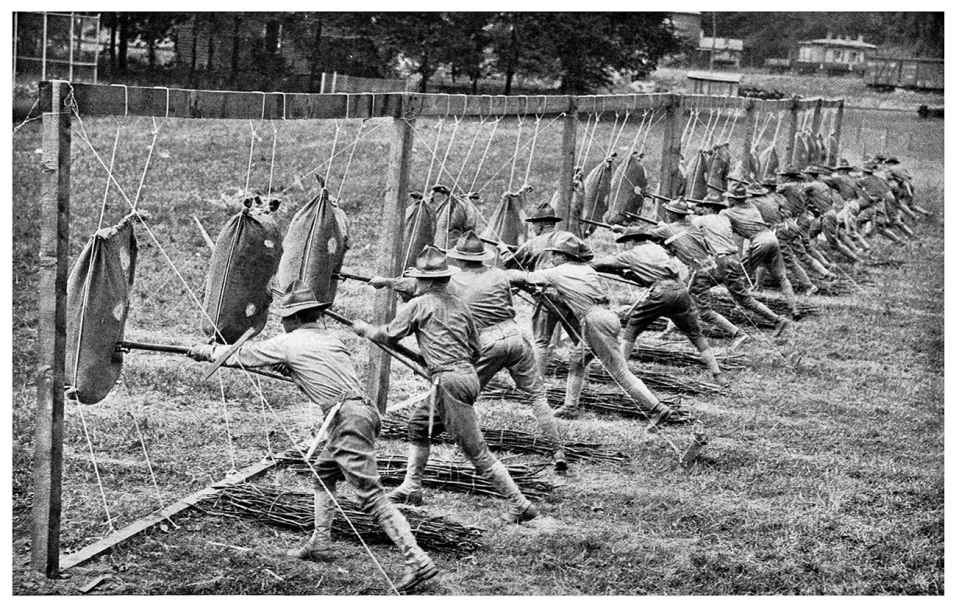 The black and white photograph shows a row of soldiers inserting their bayonets into the sacks hung in front of them through ropes, tied from the top to the wooden frame. A bunch of tied wood is lying next to each of the soldiers.