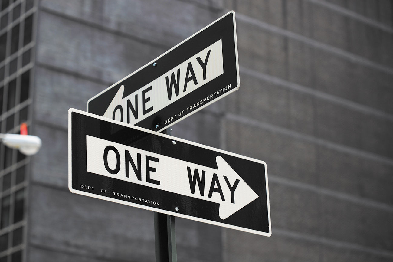 A black and white photograph of two road signs painted in the shape of arrows that reads ‘ONE WAY’, directing in different directions.