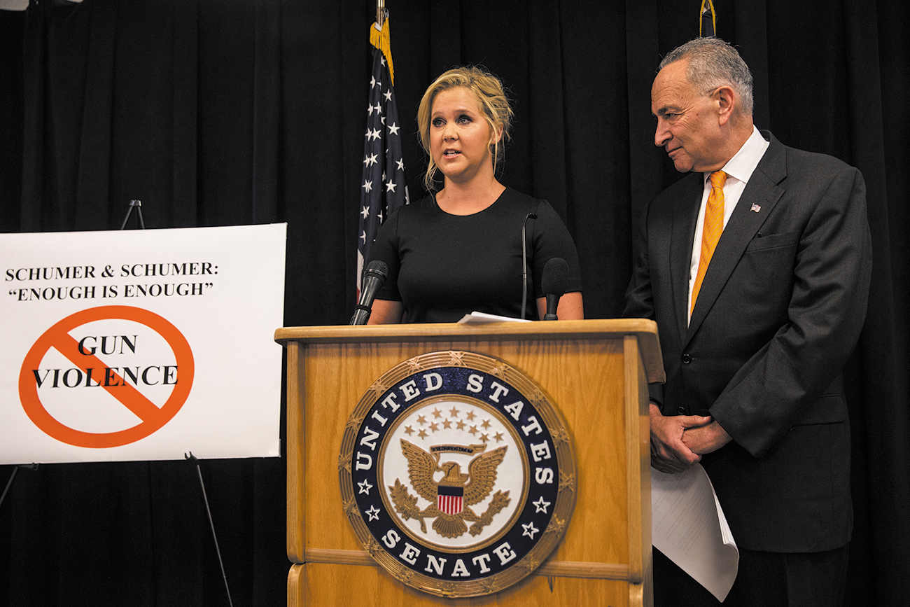 A photograph of actor Amy Schumer standing and speaking on microphone on a podium. She is Standing next to her cousin, Senator Charles Schumer. On the podium, there is an emblem, on which reads, ‘United States Senate’. On the left side of Amy Schumer, there is a flag of United States of America, and a white board which reads, ‘SCHUMER & SCHUMER: “ENOUGH IS ENOUGH” and a symbol of prohibition of gun violence.