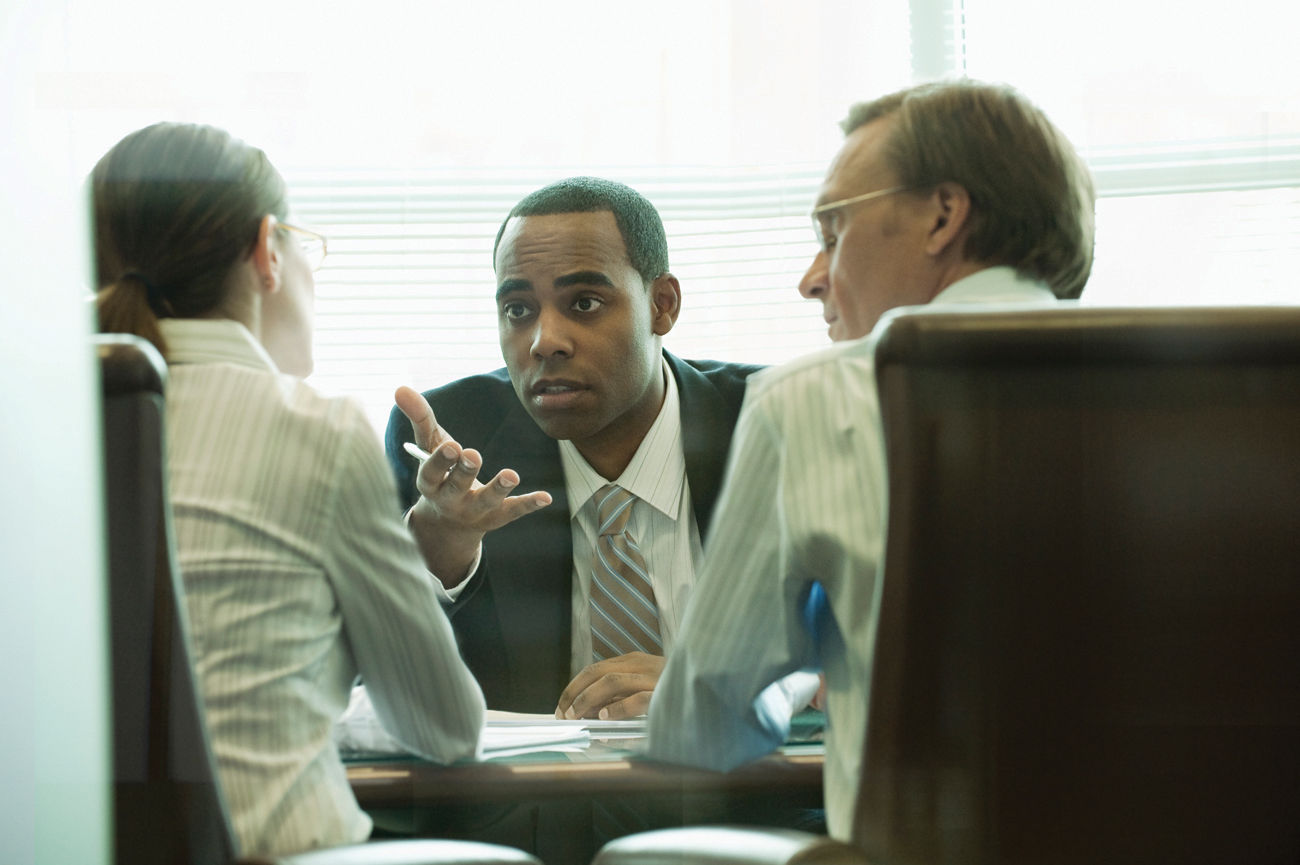 The photograph shows a man sitting in front of a woman and a man on a desk, where he is talking to the woman and making a hand gesture while making the conversation. The other man is also looking at the woman on his side.