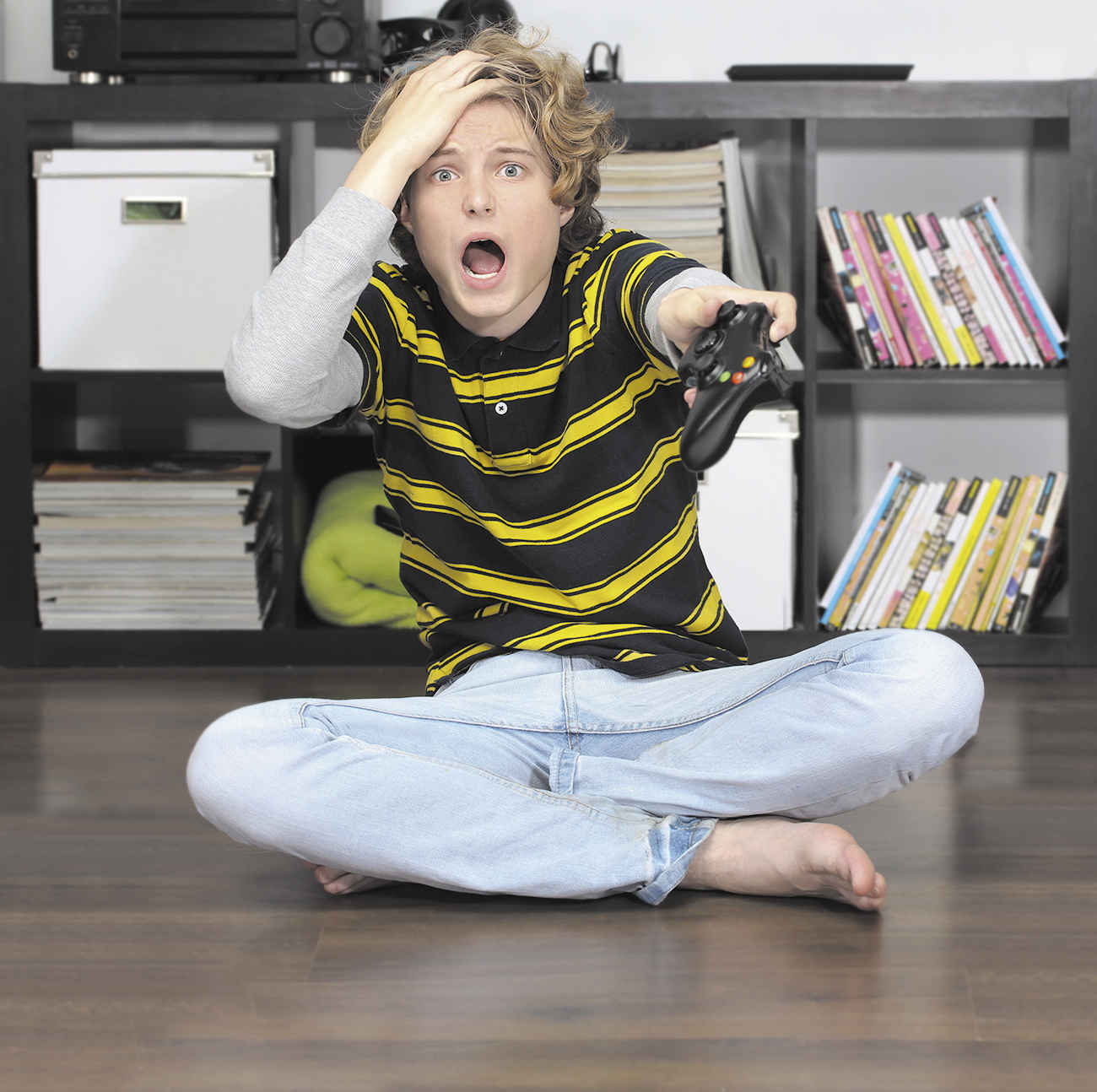 The photograph shows a boy sitting on the floor in a room looking tensed with his mouth open and eyes tensed. He is holding his head in his right hand, and a gaming remote control in his other hand, pointing towards something in the distance. A book shelf is seen behind him.