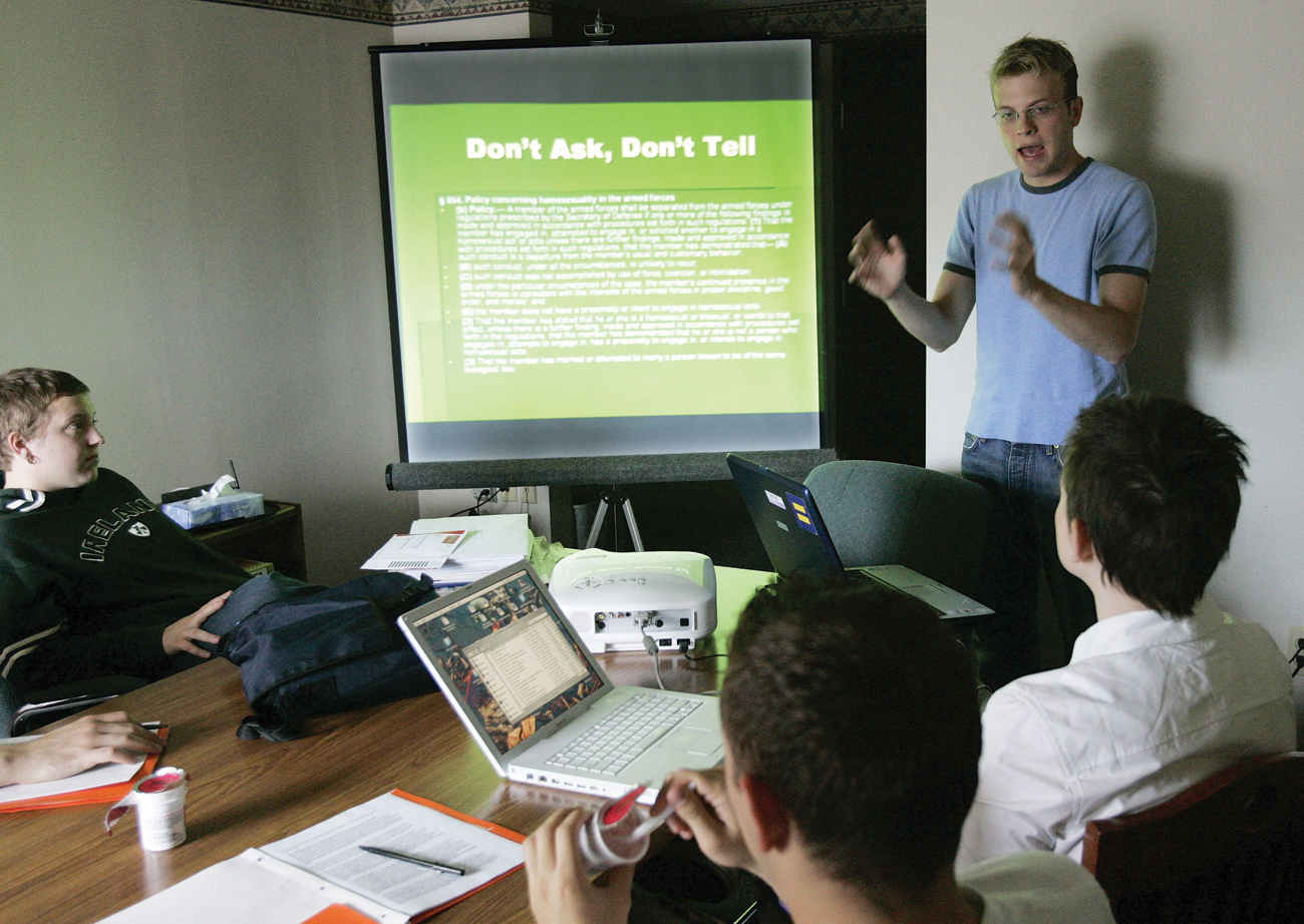 The photograph shows a man standing and giving a power point presentation on a white screen with the help of a projector. He is speaking to a group of people who are sitting in front of him, who are listening to the man speaking, and have their laptops before them, open on the table.