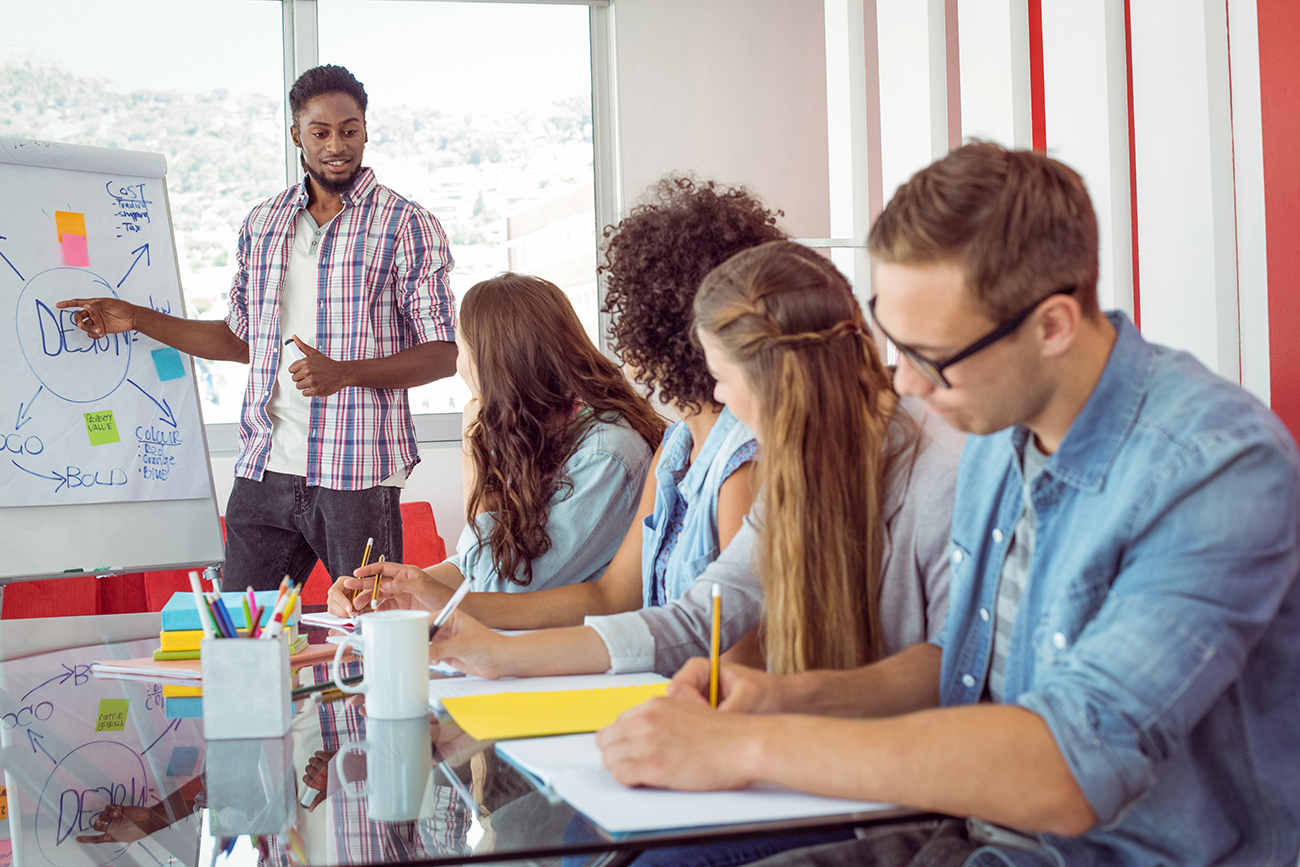 The photograph shows a man standing and giving a presentation on a white board which has a diagram on it. There are three women and a man sitting in a row on a table and chairs. While the women are looking towards the man giving presentation, the sitting man is writing something on a notebook. There are a few pens and pencils in pen holders and other stationary items on the table.