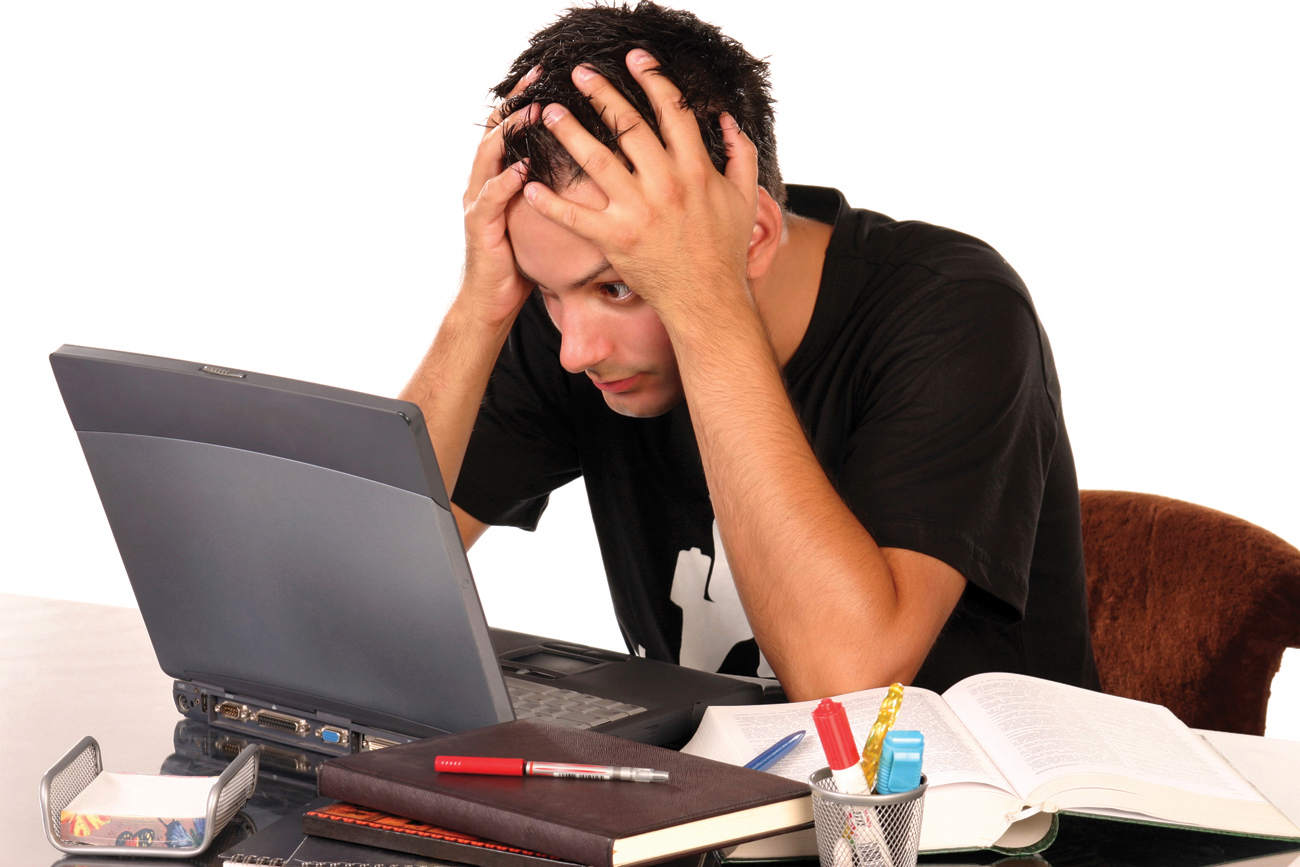 The photograph shows a man sitting in front of his laptop, kept at a table. He is holding his head in both of his hands. The table has diaries and stationary.