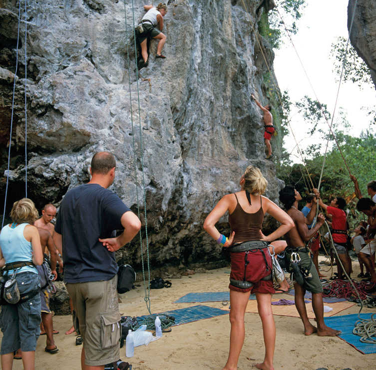 A photograph of two people doing mountaineering and several others are looking at them. Some other people are standing at the base of the mountain and looking at the two mountaineers. Some people are also preparing to start mountaineering.