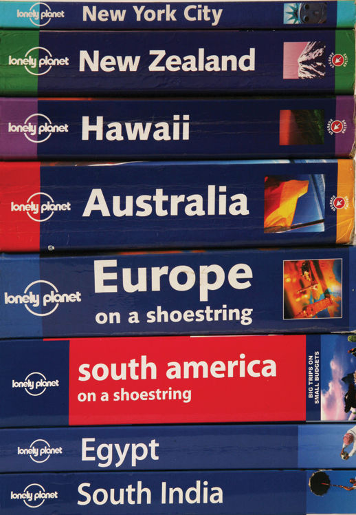 A photograph that shows books stacked one on another. The titles of the books are, ‘Lonely Planet’ followed by the country’s name they are written about, such as, ‘New York City’, ‘New Zealand’, ‘Hawaii’, ‘Australia’, ‘Europe on a shoestring’, ‘South America on a shoestring’, ‘Egypt’, ‘South India’ in a top to bottom order