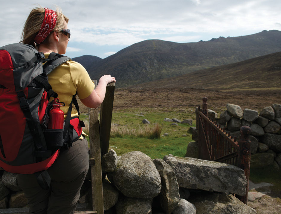 The photograph shows back side of a woman who is wearing a bag pack, sunglasses and a head band. There are small rocks around her feet and she is holding a wooden stand. She is looking at the mountains in the distance 