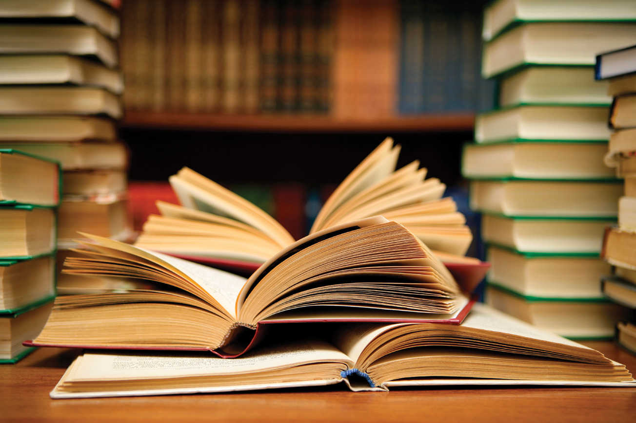 The photograph shows three open books lying one above other on the middle of a table and on the left and right of this a pile of books are kept. A long book shelf can be seen the background.