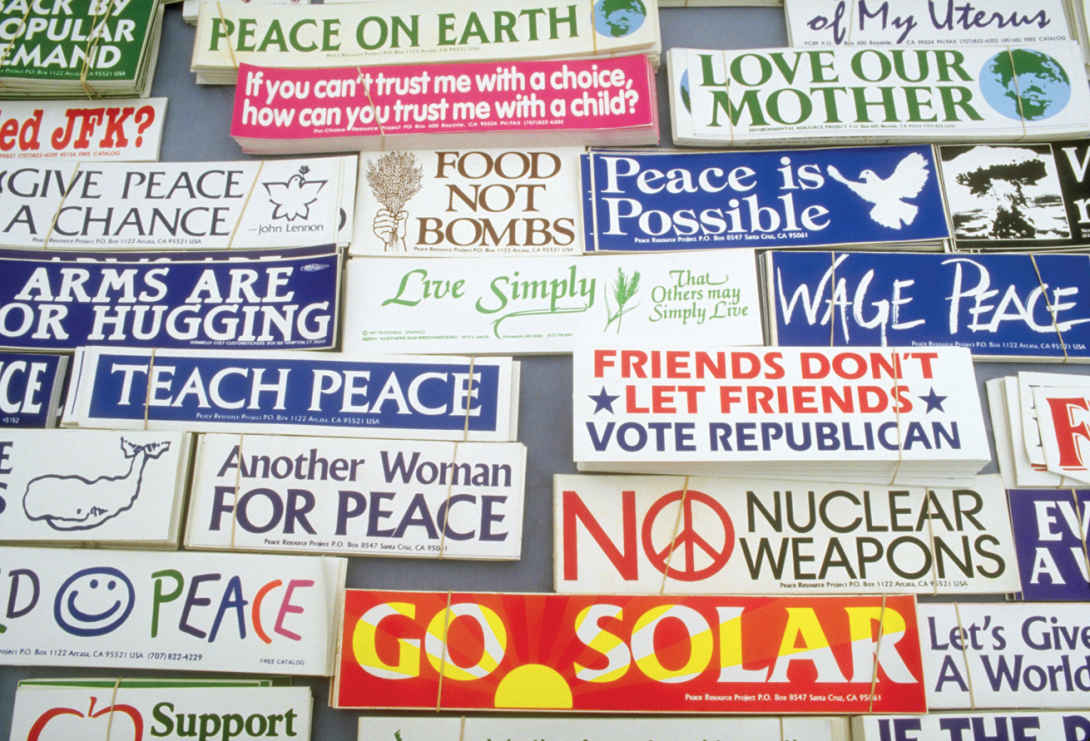 Image shows a lot of bumper stickers with different slogans, but with the common theme of peace. Some of the slogans read, “Peace on Earth”, “Arms are for Hugging”, “Teach Peace”, “No Nuclear Weapons”, “Go Solar”, and “Peace is possible”, among others.