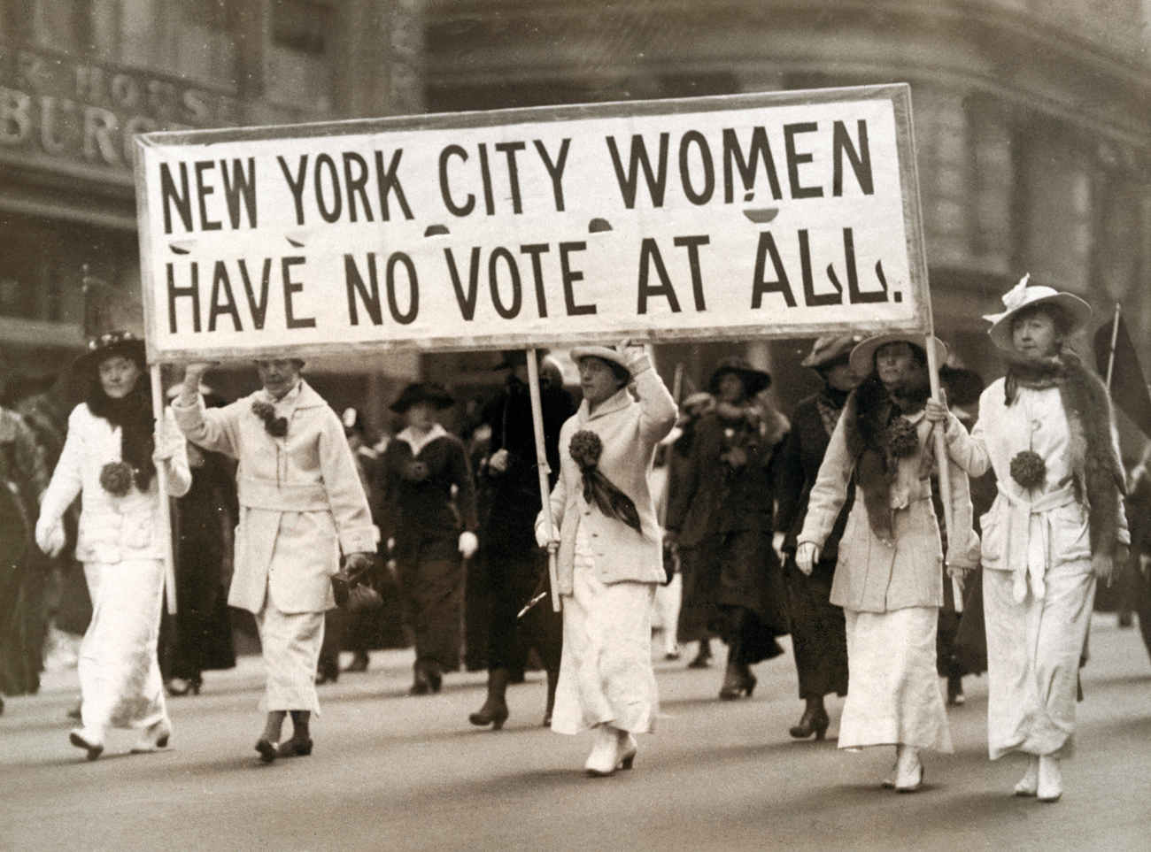 A photograph (dated 1913) of a group of women marching on a New York street demanding voting rights. They are led by 4 women dressed in white clothes who are holding a banner that reads “New York City women have no vote at all.” They all are wearing scarves, winter coats, long skirts and boots and their dresses reflect affluence.