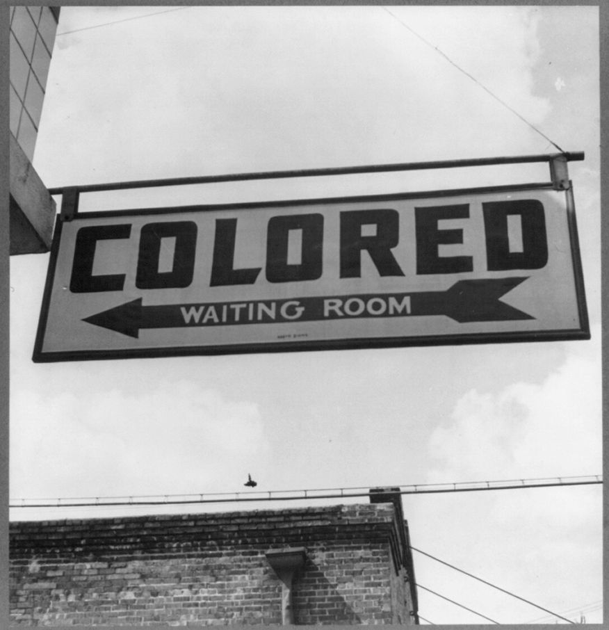 A signboard that points towards a seperate waiting room for colored people. The word “colored” is written in a large font and words “Waiting Room” are written in a smaller font onto an arrow that points towards the left.