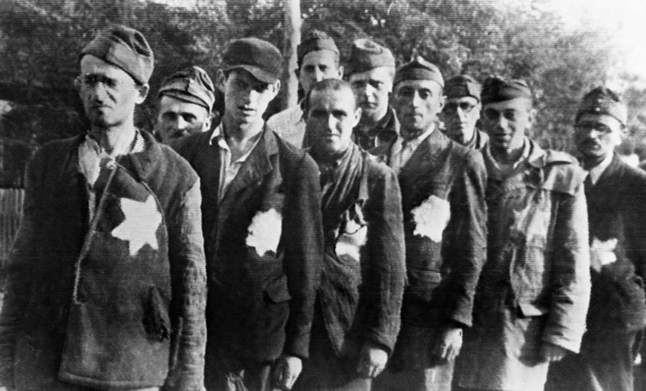 A photograph depicting 10 men lined up behind one another wearing coats that have the “Star of David” emblazoned on the left side of the coat. They are of varying age-groups and are wearing old and ill-fitted coats and caps on their heads.